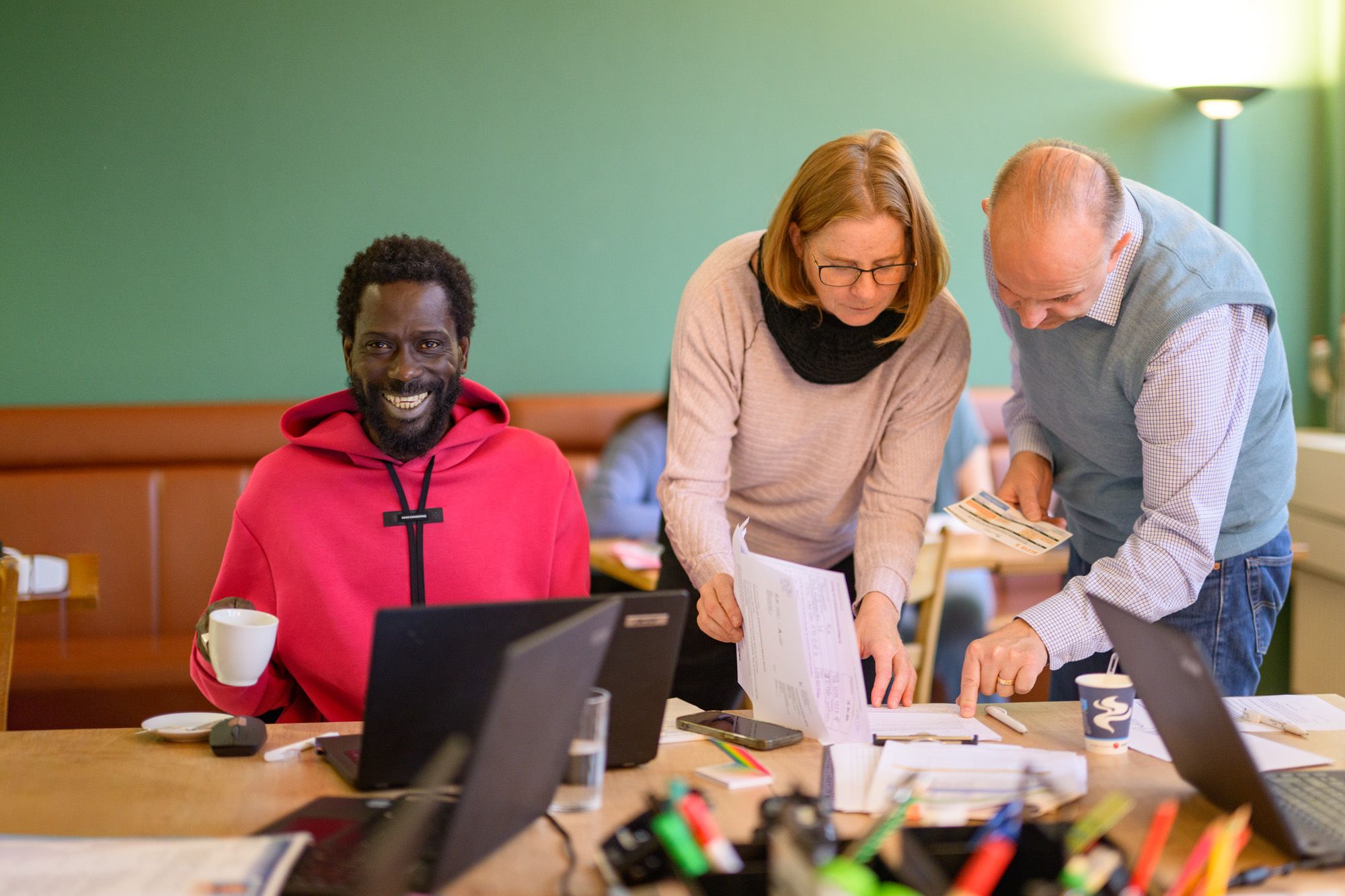 Ein Besuchender der Lernstube lächelt in die Kamera, er hat einen Kaffee in der Hand. Neben ihm sprechen zwei Lernbegleitungen miteinander.