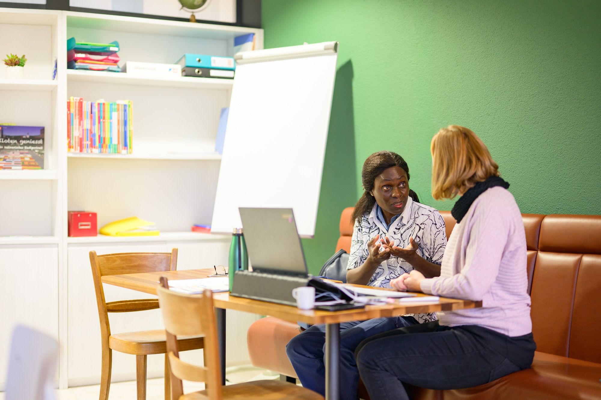 Eine Besuchende der Lernstube Kloten ist im Gespräch mit einer Lernbegleitung, welche ihr am Laptop etwas erklärt. Die beiden sitzen auf einer gemütlichen, braunen Sitzbank