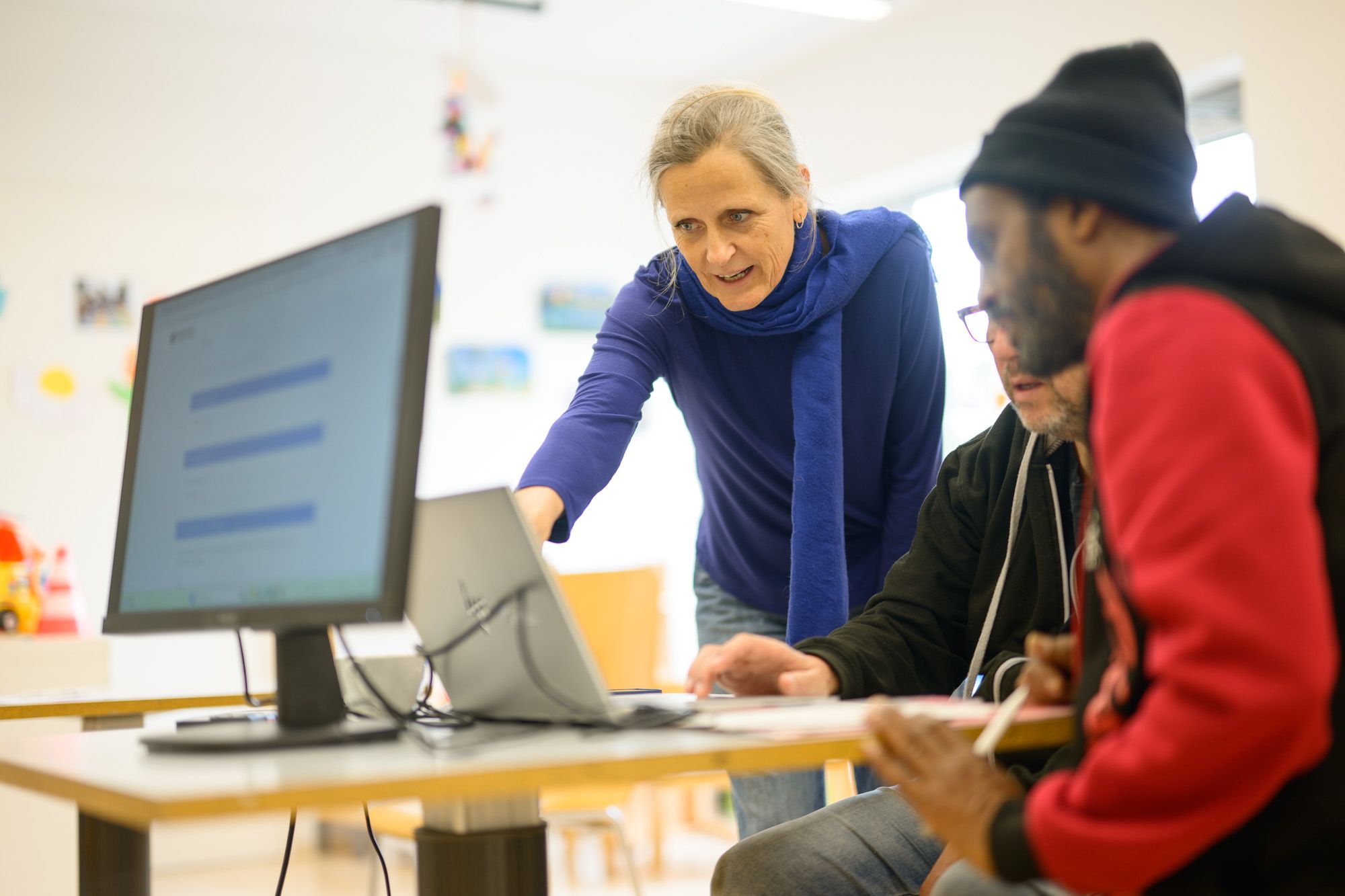 Bild der Lernstuben-Leitung Linda Gosteli, die zwei Personen an einem Laptop etwas erklärt.